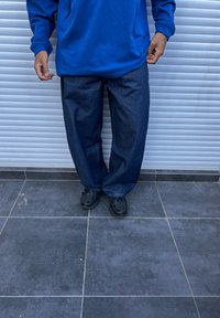 Sinine pikaaärisega särk, mis on sobitatud laia tumedate teksadega. Mustad spordijalatsid täiendavad komplekti, taustal on hall plaatidega pind.