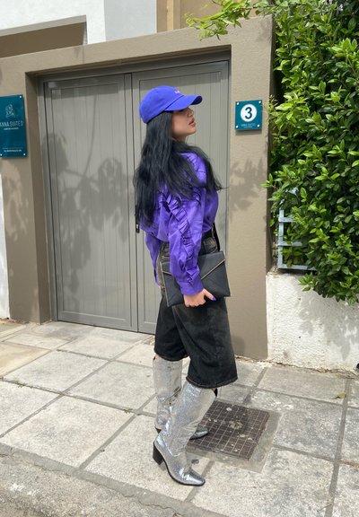 Camisa de manga larga morada con mangas acampanadas, pantalones cortos negros de talle alto y botas plateadas hasta la rodilla con un acabado brillante. Sosteniendo un bolso de mano negro.