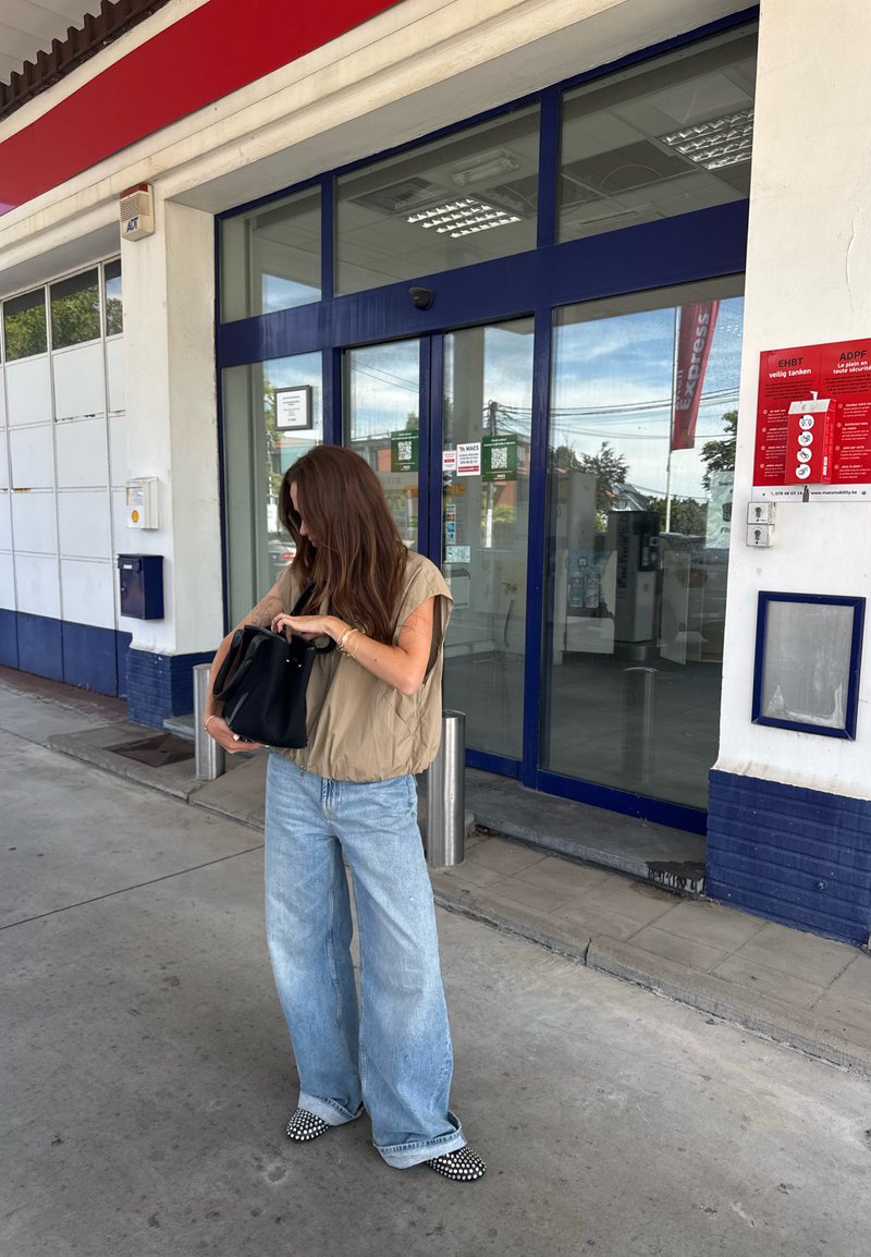 Blusa corta bege de mangas curtas, jeans largos azul claro, sapatos pretos com padrão xadrez, mala preta. Cenário ao ar livre com portas de vidro e sinalização visível.