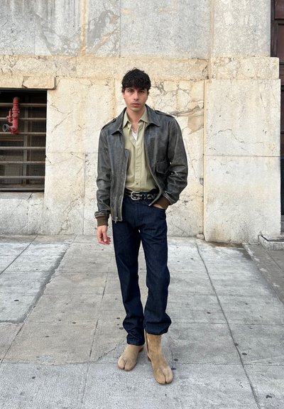 Chaqueta de cuero gris, camisa de oliva abotonada, jeans azul oscuro y botas de tobillo beige. De pie contra una pared de piedra texturizada.