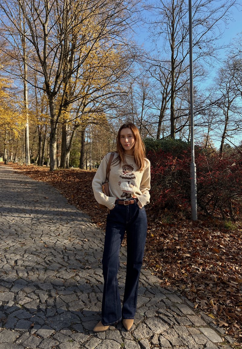 Pull crème avec un imprimé, jean évasé bleu foncé, ceinture en cuir marron et chaussures à talons beige, dans un parc aux feuillages d'automne.
