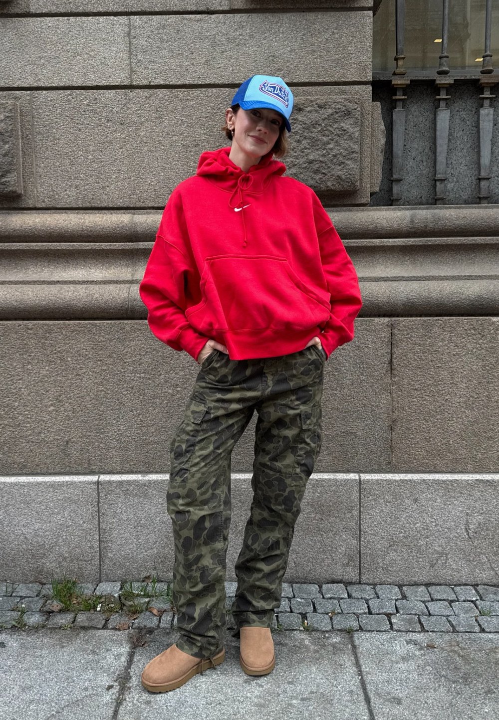 Sudadera roja con capucha y bolsillo frontal, pantalones cargo de camuflaje, botas marrones y una gorra azul con logotipo blanco. Entorno urbano al aire libre.