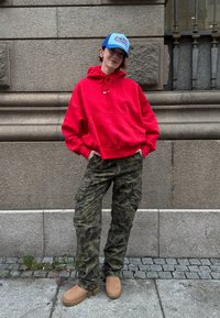 Sudadera roja con capucha y bolsillo frontal, pantalones cargo de camuflaje, botas marrones y una gorra azul con logotipo blanco. Entorno urbano al aire libre.