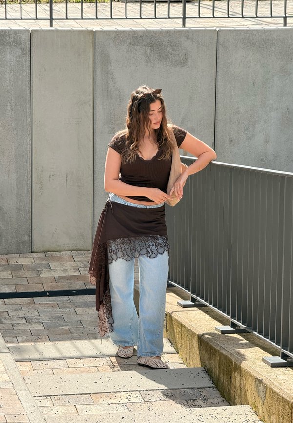 Kvinde i sort top og blonder omslagsnederdel over jeans justerer taskestrop, mens hun står ved metalgelænder på en solrig brolagt sti.