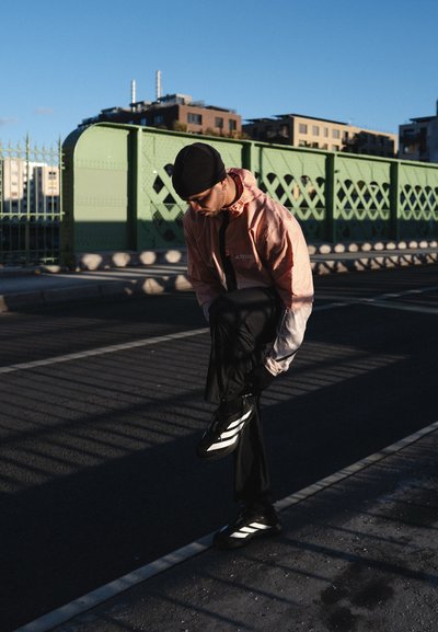Hombre con gorro negro y chaqueta color melocotón atándose las zapatillas negras con rayas blancas en una calle urbana cerca de una valla metálica verde al atardecer.