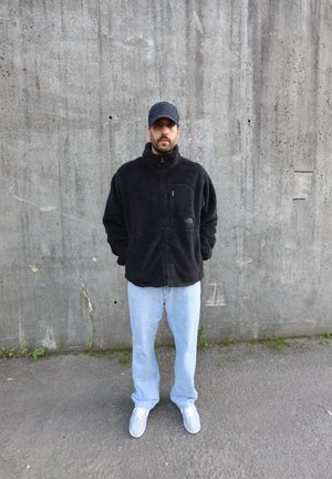 Chaqueta de forro polar negra con cuello alto y bolsillo con cremallera, combinada con jeans azul claro y zapatillas grises, frente a una pared de concreto.