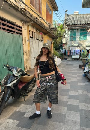Joven mujer con pantalones cortos de camuflaje y top negro de manga corta de pie en una calle con baldosas, con motocicletas y edificios antiguos al fondo.