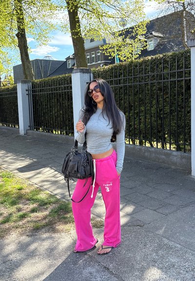Mujer con camiseta de manga larga gris, pantalones deportivos rosa brillante y gafas de sol, de pie en la acera sosteniendo un bolso negro cerca de una valla negra.