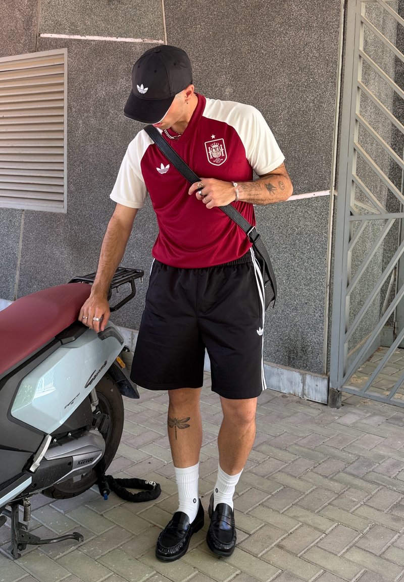 Un homme portant un maillot de sport rouge et blanc et un short noir se tient à côté d'un scooter, ajustant la sangle de son sac d'épaule noir.