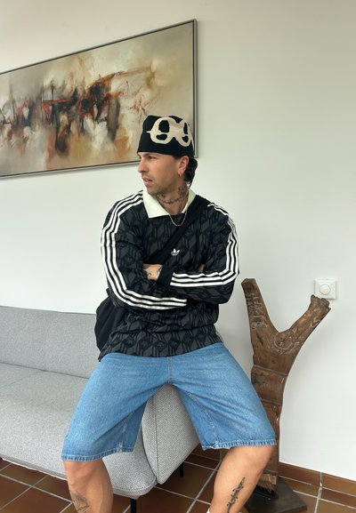 Camisa negra con estampado y mangas largas, con rayas blancas, pantalones cortos de denim azul claro y un gorro blanco y negro, sentado en un sofá gris.