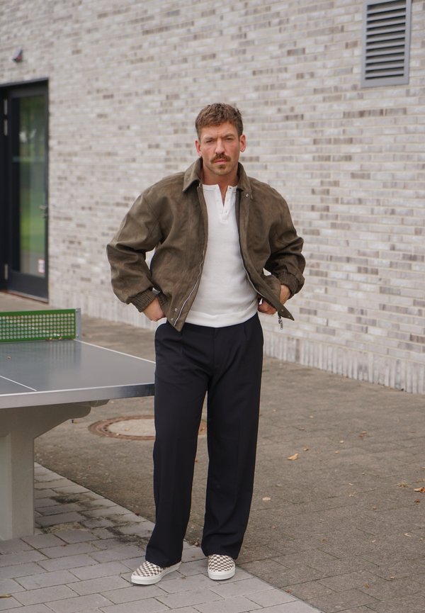Hombre con chaqueta marrón, camisa blanca, pantalones negros y zapatos tipo slip-on a cuadros, de pie con las manos en los bolsillos cerca de una mesa de ping pong exterior.