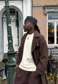 Casaco oversized castanho sobre um suéter claro com o texto "QUOTED", combinado com calças castanhas, joias douradas e um boné preto. O fundo inclui um poste de luz e uma bicicleta.