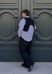 Homme en pantalon noir, gilet noir sur une chemise blanche, et chaussures noires, debout de profil devant une grande porte verte ornée.