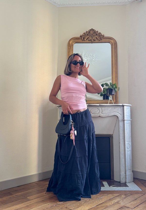 Saia maxi preta em camadas com tecido texturizado, blusa justa rosa claro e uma pequena bolsa preta. Inclui óculos de sol redondos e charmosos adornos decorativos.