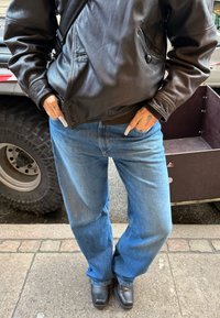 Personne portant une veste en cuir noire, un jean bleu ample, des chaussures noires à bout carré, les mains dans les poches et des tatouages visibles sur les doigts.