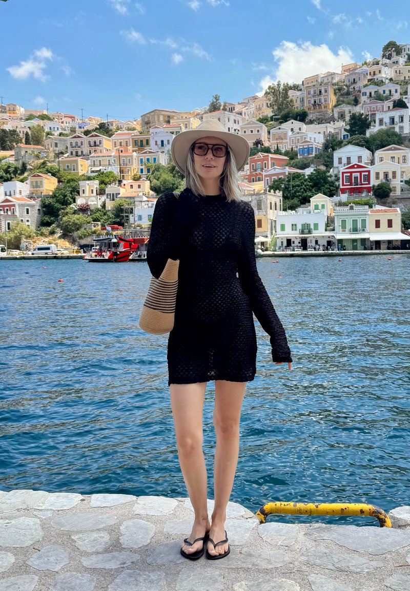 Femme en robe noire et chapeau debout sur un quai en pierre au bord d’une eau bleue avec des maisons colorées sur la colline et un ciel clair en arrière-plan.