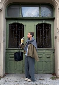 Chaqueta de algodón oversize verde oliva, bufanda gris, camisa blanca, pantalones anchos grises, bolso tote negro, zapatos beige, frente a una puerta verde.