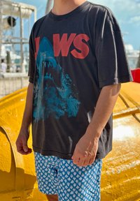 Personne portant un t-shirt noir JAWS avec un motif de requin et un short bleu à motifs, debout à l'extérieur près d'une grande structure jaune.