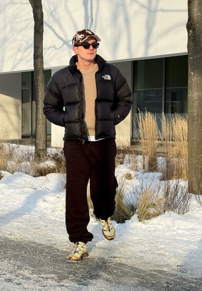 Hombre con chaqueta acolchada negra, pantalones marrones, gorra de camuflaje y gafas de sol caminando por un camino nevado cerca de árboles y hierba seca.