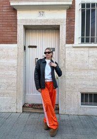 Μαύρο δερμάτινο μπουφάν, λευκή μπλούζα, πορτοκαλί φόρμα με λευκές ρίγες, καφέ μπότες και γυαλιά ηλίου, στέκεται δίπλα σε μια πόρτα με τον αριθμό 57.