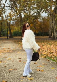 Chlpatá biela umelá kožušina, sivé rozšírené nohavice, svetlé tenisky a čierna kabelka. Pozadie tvorí jesenné lístie a chodník.