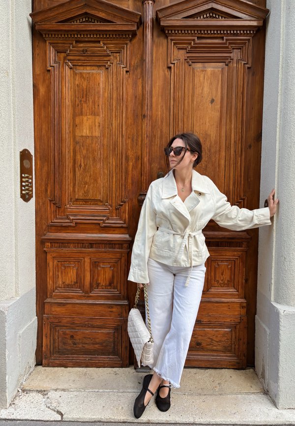 Donna con giacca e pantaloni bianchi che tiene una borsa trapuntata, appoggiata a una porta in legno con intagli intricati e pannello citofono a sinistra.