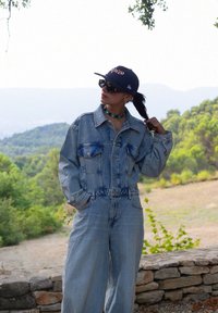 Hele sinine denim kombinesoon nööbidest ees ja suurte taskutega, kombineeritud tumesinise mütsi ja mustade prillidega, roheline taust.
