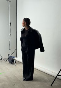 Schwarze, kurze Lederjacke über einem schwarzen, durchsichtigen Oberteil und lockeren schwarzen Hosen. Das Modell steht vor einem schlichten Hintergrund und ist seitlich zu sehen.