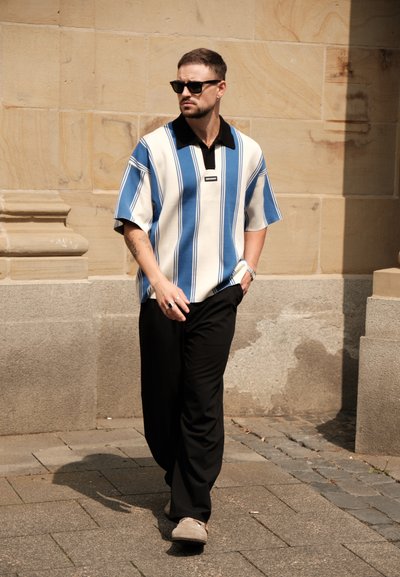 Camisa de manga corta con rayas azules y blancas y un cuello negro; combinada con pantalones negros y zapatos grises, de pie sobre un pavimento de piedra.
