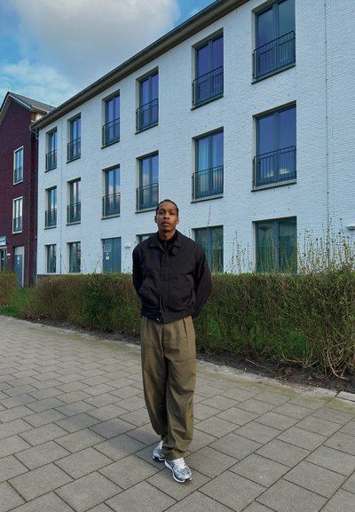 Una persona está de pie en un camino pavimentado, vestida con una chaqueta negra, pantalones verdes y zapatillas plateadas, con un edificio de ladrillo y vegetación al fondo.