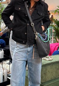 Borsa a tracolla nera con una tracolla intrecciata blu e verde, hardware argentato e un design elegante. Indossata con jeans di denim azzurri e una giacca nera.