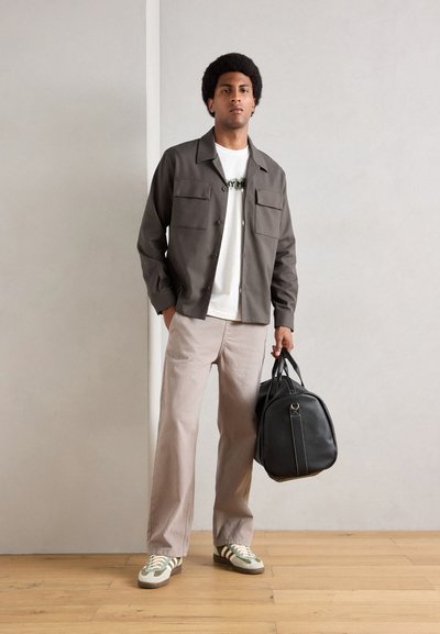 Chaqueta de botones gris sobre una camisa blanca, pantalones beige y bolso negro. Zapatillas texturizadas con rayas verdes y blancas. Fondo de suelo de madera.