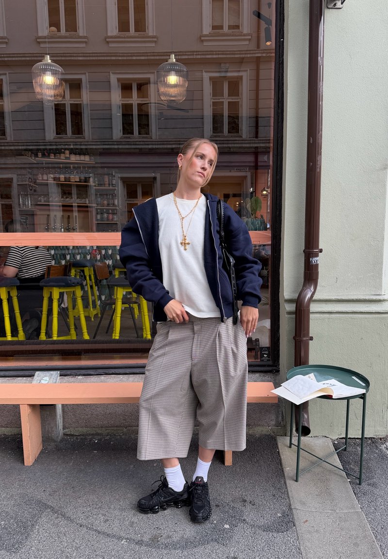 Jeune femme en veste bleu marine et pantalon large à carreaux appuyée sur un banc couleur pêche devant un café avec une grande fenêtre et des tabourets jaunes à l'intérieur.