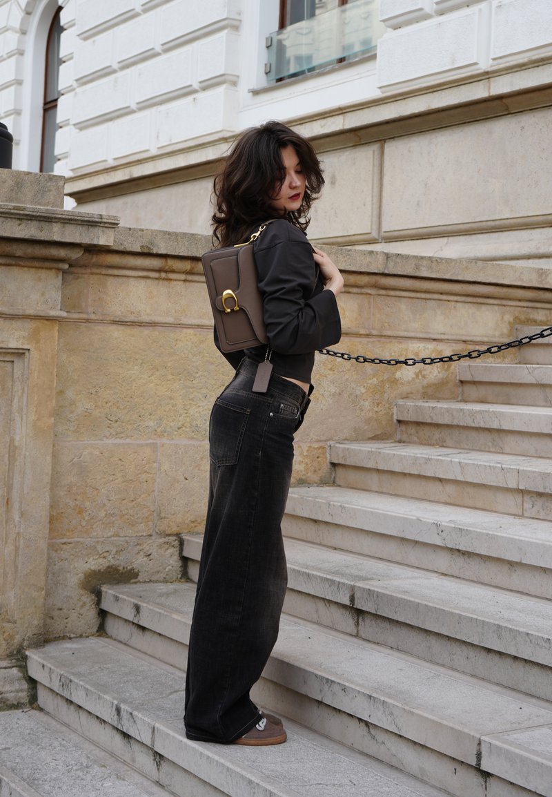 Joven mujer con cabello oscuro, vestida con top negro y vaqueros, llevando un bolso marrón, de pie en unas escaleras de piedra al aire libre junto a un edificio histórico.