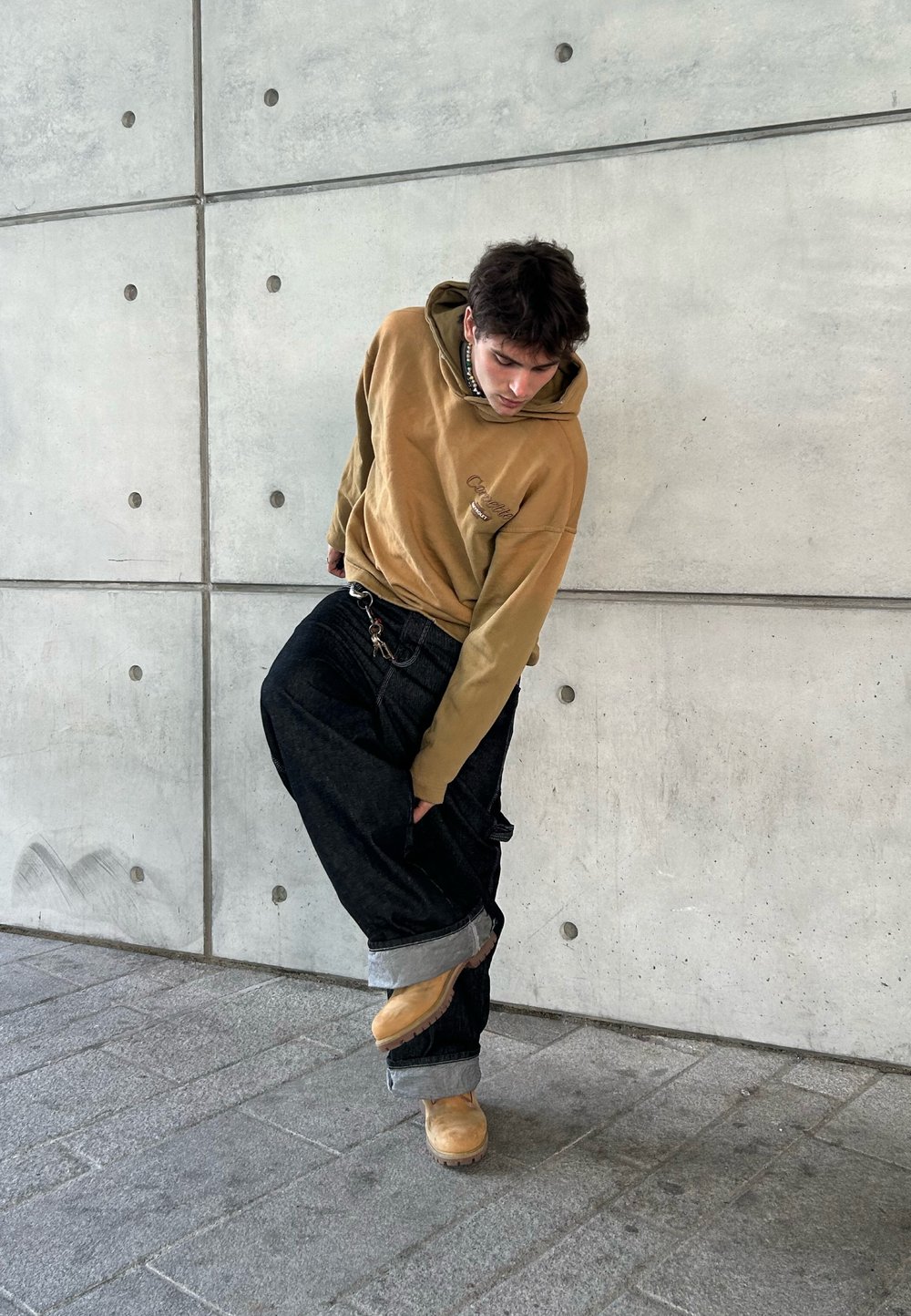 Hoodie castanho com um logótipo, calças de ganga pretas largas com a bainha rolada e tamancos bejes. Posicionado contra uma parede de betão com parafusos arredondados.