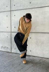 Hoodie castanho com um logótipo, calças de ganga pretas largas com a bainha rolada e tamancos bejes. Posicionado contra uma parede de betão com parafusos arredondados.