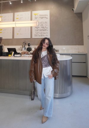 Mujer con cabello largo, chaqueta de cuero marrón, camisa blanca y vaqueros azules, de pie dentro de una cafetería moderna cerca del mostrador con el menú en la pared.