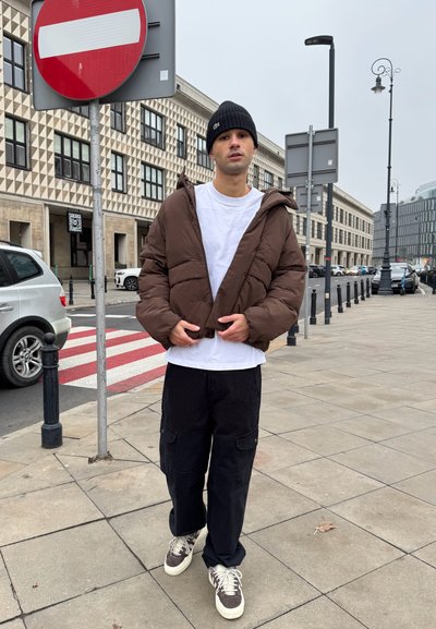 Chaqueta de plumas marrón con capucha, camiseta blanca y pantalones cargo negros. Zapatillas grises, entorno urbano con señales de tráfico y edificios al fondo.