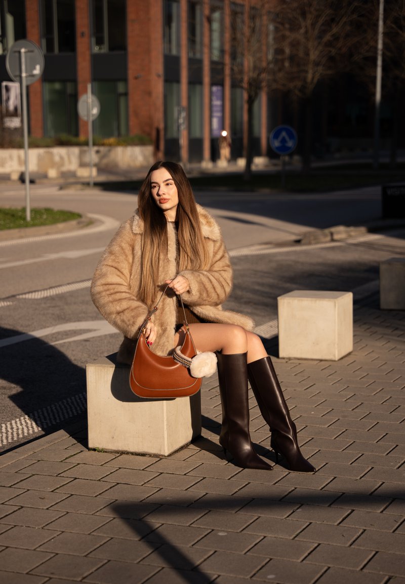Young woman in beige fur coat and black knee-high boots sitting on a concrete block, holding a brown handbag on a city sidewalk.