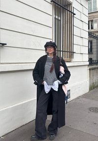 Femme avec une longue tresse, lunettes, casquette noire, pull gris, chemise rayée, manteau noir, pantalon large, debout sur le trottoir près d'un bâtiment blanc avec une fenêtre grillagée.