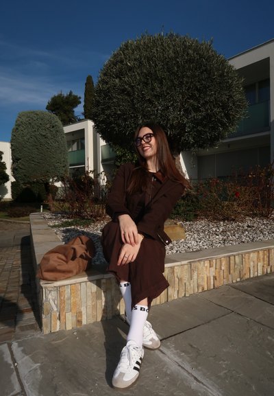 Mujer con cabello largo y castaño y gafas sentada en un borde de piedra junto a arbustos, vestida con un conjunto marrón, zapatillas blancas y sonriendo al aire libre en un día soleado.