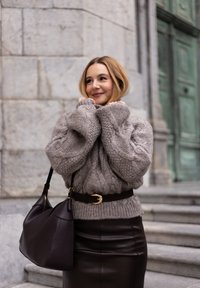 Gruby szary sweter dzianinowy z kabelkowym wzorem, czarna skórzana spódnica, brązowy pasek z złotą klamrą oraz ciemna torebka. W tle schody i kamienna ściana.