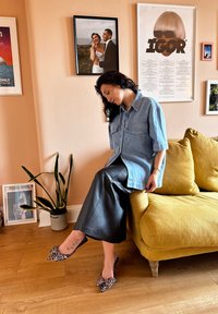 Camisa de ganga oversized com mangas curtas, dois bolsos frontais; calças de pele largas em preto mate; sapatos slip-on com estampado de leopardo. Sofá amarelo ao fundo.