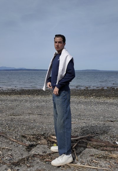 Joven con sudadera azul marino y vaqueros está de pie en una playa rocosa con océano y colinas lejanas bajo un cielo nublado.