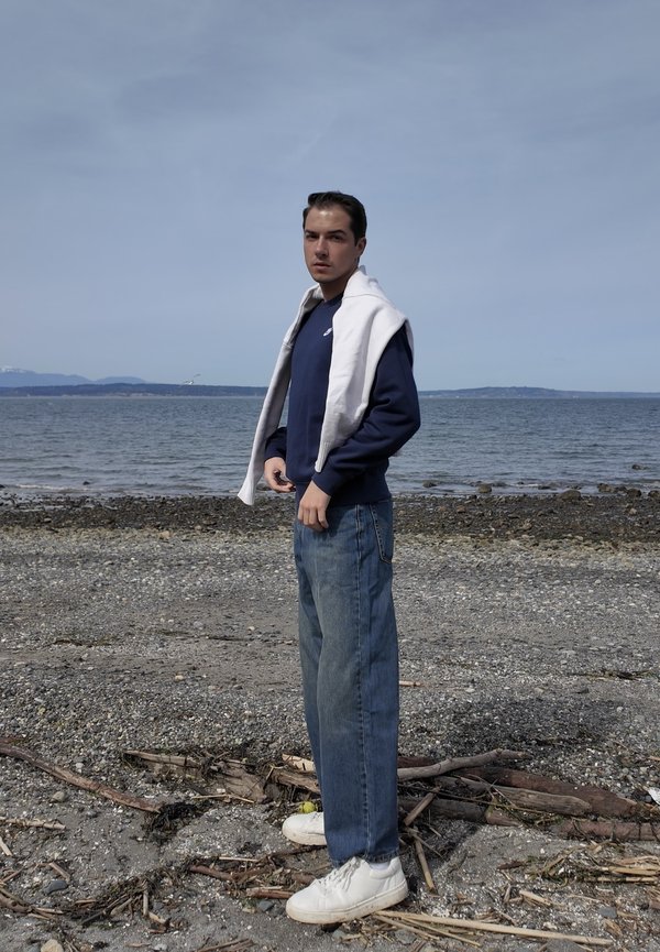 Młody mężczyzna w granatowej bluzie i dżinsach stoi na skalistej plaży z oceanem i odległymi wzgórzami pod zachmurzonym niebem.
