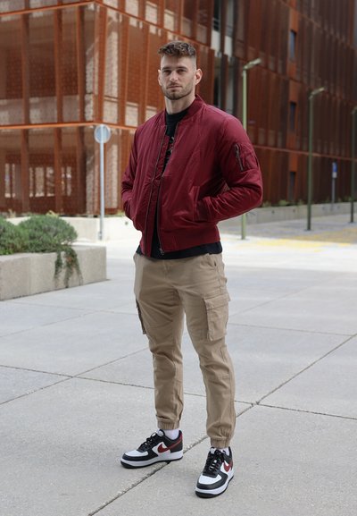 Chaqueta bomber color burdeos con bolsillos con cremallera, pantalones cargo color caqui y zapatillas Nike en blanco y negro. Textura suave y diseño ajustado.