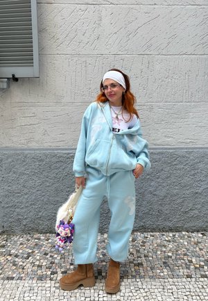 Sudadera con cremallera de color azul claro con diseño blanco, pantalones de chándal a juego; botas de plataforma color beige; diadema blanca; llevando un bolso suave y un juguete de peluche.