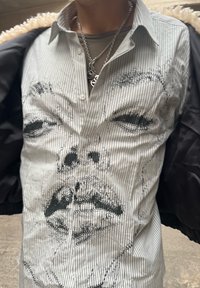 Koszula w paski z czarnym haftowanym wzorem twarzy. Tkanina wydaje się lekka, z kołnierzykiem i guzikami.