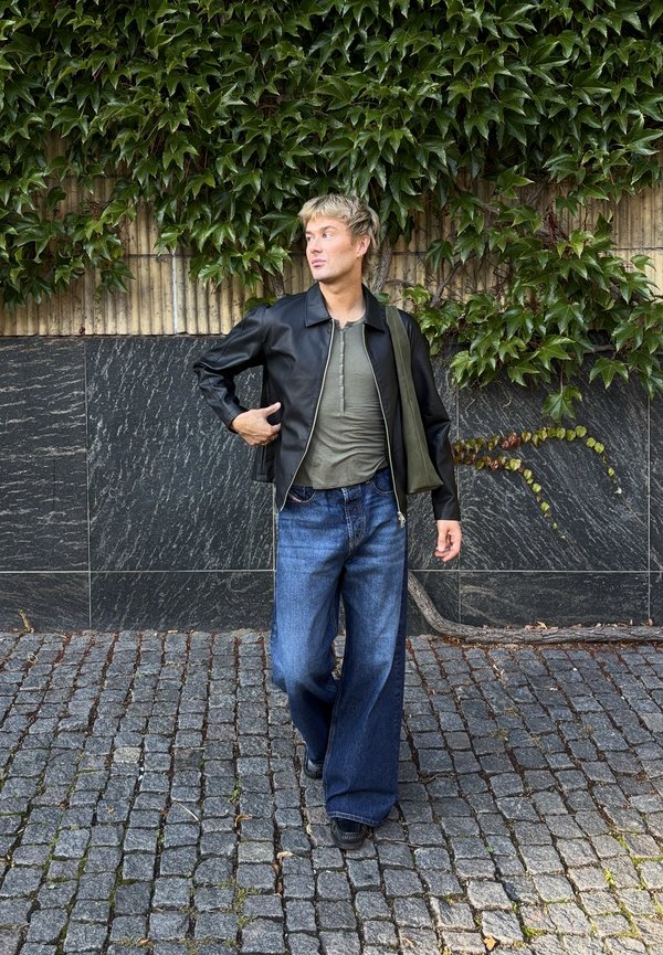 Un hombre con una chaqueta de cuero negra, una camisa gris y unos vaqueros holgados está de pie en una calle empedrada junto a una pared cubierta de hiedra.
