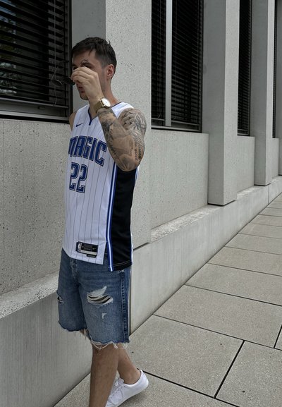 Camiseta de baloncesto blanca con acentos en azul y negro, con rayas verticales y el número 22. Combinada con pantalones cortos de mezclilla desgastados y zapatillas blancas.